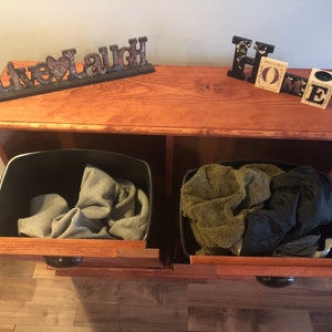 K&ouml;nnte beinhalten: Ein Holzschr&auml;nkchen mit zwei offenen Schubladen, jede mit einem schwarzen Beh&auml;lter gef&uuml;llt mit Kleidung. Der Schrank hat eine Holzplatte mit den Worten "Live Laugh" und "Home" darauf.