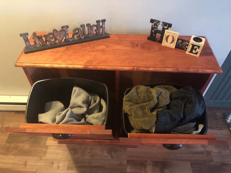 K&ouml;nnte beinhalten: Ein Holzschr&auml;nkchen mit zwei offenen Schubladen, jede mit einem schwarzen Beh&auml;lter gef&uuml;llt mit Kleidung. Der Schrank hat eine Holzplatte mit den Worten "Live Laugh" und "Home" darauf.