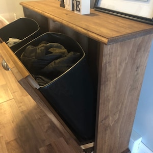 K&ouml;nnte beinhalten: Ein Holzaufbewahrungsschrank mit zwei herausziehbaren Beh&auml;ltern. Die Beh&auml;lter sind schwarz und haben einen Metallrahmen. Der Schrank hat eine braune Beize und steht auf einem Holzboden.