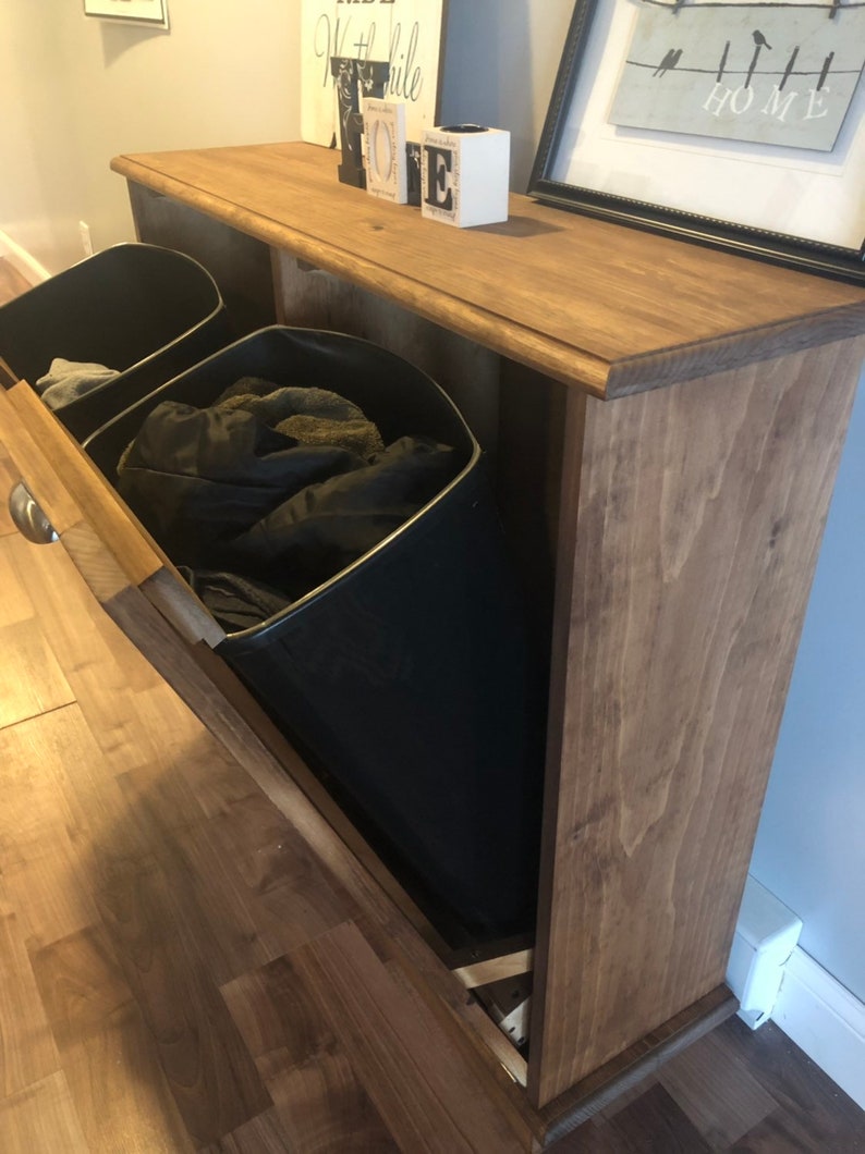 K&ouml;nnte beinhalten: Ein Holzaufbewahrungsschrank mit zwei herausziehbaren Beh&auml;ltern. Die Beh&auml;lter sind schwarz und haben einen Metallrahmen. Der Schrank hat eine braune Beize und steht auf einem Holzboden.