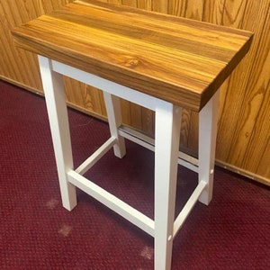 Peut inclure: Un tabouret en bois blanc avec un plateau en bois naturel. Le tabouret a un design simple avec quatre pieds et un plateau rectangulaire.