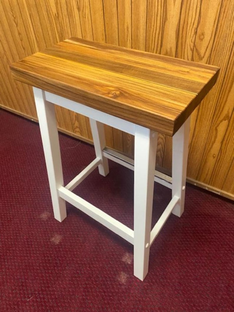 Peut inclure: Un tabouret en bois blanc avec un plateau en bois naturel. Le tabouret a un design simple avec quatre pieds et un plateau rectangulaire.