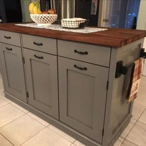 Puede incluir: Una isla de cocina gris con una encimera de madera y cuatro puertas de armario. La isla tiene herrajes negros y un toallero en el lateral.