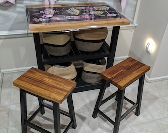 May include: Two black and wood bar stools with a black metal frame and a wooden seat. The stools are in front of a black metal kitchen cart with three shelves. The cart has a wooden top and is holding baskets.
