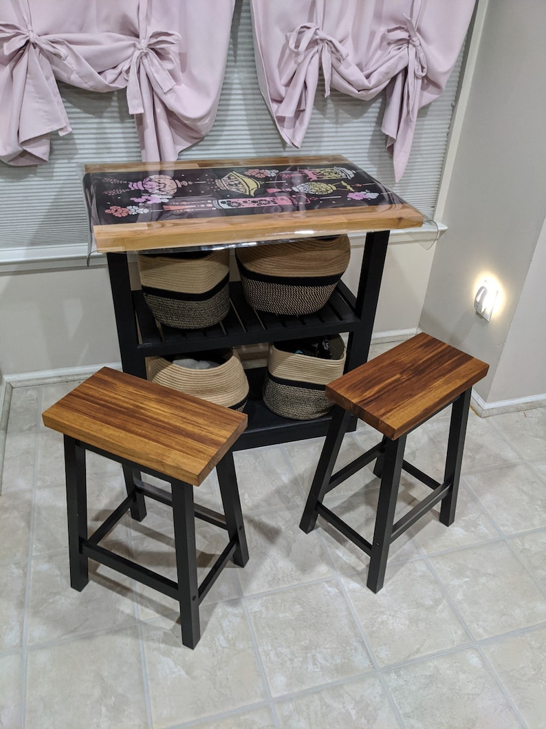 Peut inclure: Deux tabourets de bar en bois et noir avec une structure en m&eacute;tal noir et un si&egrave;ge en bois. Les tabourets sont devant un chariot de cuisine en m&eacute;tal noir avec trois &eacute;tag&egrave;res. Le chariot a un dessus en bois et contient des paniers.