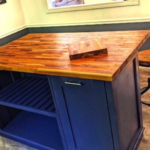 Op de afbeelding: Een blauw keukeneiland met een houten aanrechtblad en een plank. Het eiland heeft een enkele deur en een klein snijplank bovenop. Een houten kruk met een metalen basis staat naast het eiland.