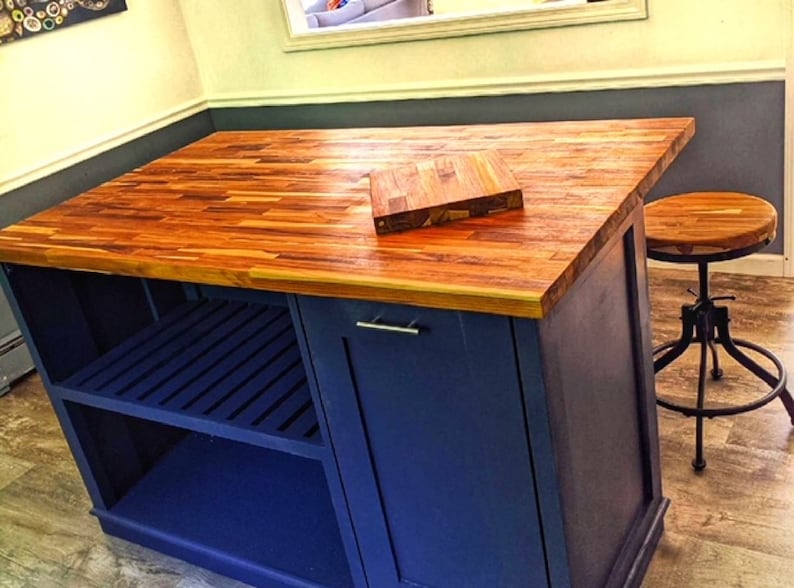 Op de afbeelding: Een blauw keukeneiland met een houten aanrechtblad en een plank. Het eiland heeft een enkele deur en een klein snijplank bovenop. Een houten kruk met een metalen basis staat naast het eiland.
