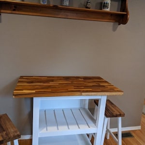 Peut inclure: &Icirc;lot de cuisine blanc avec un comptoir en bois de boucher et deux tabourets blancs. L'&icirc;lot a deux &eacute;tag&egrave;res et une base blanche.