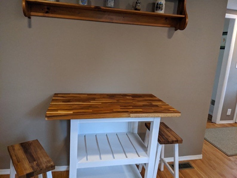 Peut inclure: &Icirc;lot de cuisine blanc avec un comptoir en bois de boucher et deux tabourets blancs. L'&icirc;lot a deux &eacute;tag&egrave;res et une base blanche.