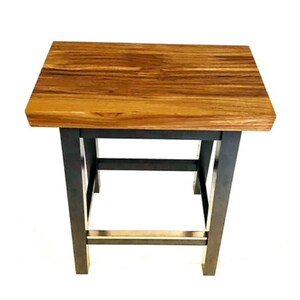 Peut inclure: Un tabouret en bois avec un plateau en bois naturel et des pieds en m&eacute;tal noir. Le tabouret a un design simple avec un plateau rectangulaire et une base carr&eacute;e.