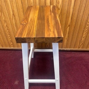 Peut inclure: Un tabouret en bois avec une structure peinte en blanc et un plateau en bois naturel. Le tabouret a un design simple avec un plateau rectangulaire et quatre pieds.