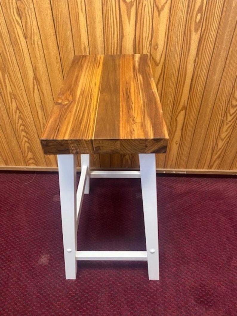 Peut inclure: Un tabouret en bois avec une structure peinte en blanc et un plateau en bois naturel. Le tabouret a un design simple avec un plateau rectangulaire et quatre pieds.