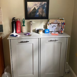K&ouml;nnte beinhalten: Wei&szlig;er Schrank mit zwei T&uuml;ren und zwei ausziehbaren M&uuml;lleimern. Der Schrank hat silberne Griffe und steht auf einem wei&szlig;en Sockel. Die Oberseite des Schranks ist mit einem gerahmten Foto, einer gr&uuml;nen Fusselrolle, einer roten und einer schwarzen Wasserflasche, einem wei&szlig;en Zylinder, einem kleinen wei&szlig;en Lautsprecher, einer Kerze und einem blauen JBL-Lautsprecher dekoriert.