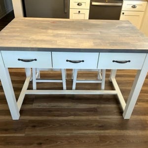 May include: White kitchen island with a gray countertop and three drawers with black handles. The island has a white base and is sitting on a wood floor.