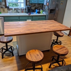 Op de afbeelding: Een wit keukeneiland met een houten aanrechtblad en vier zwarte metalen barstoelen met houten zittingen. Het eiland bevindt zich in een keuken met groene kasten en een koelkast van roestvrij staal.