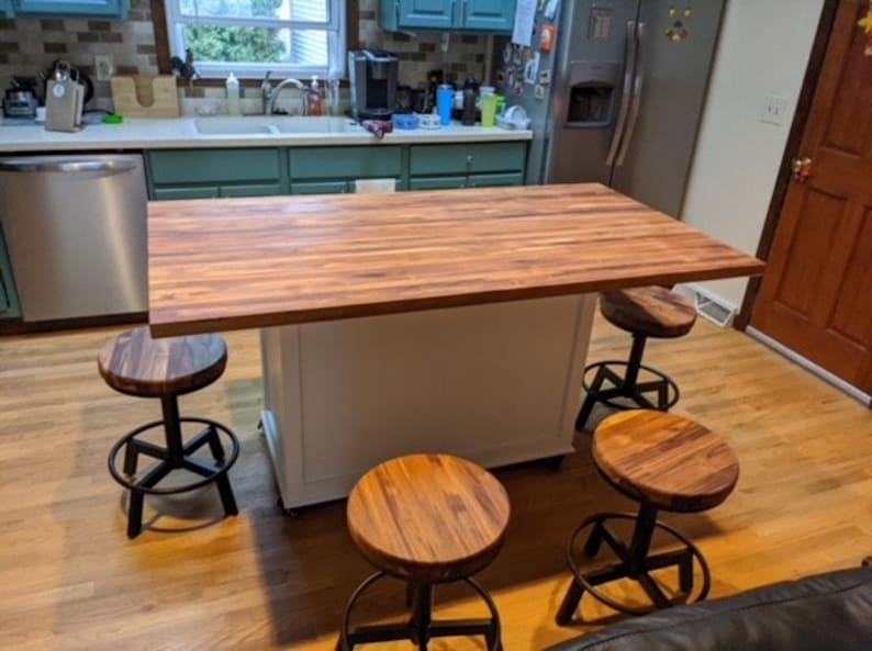 Op de afbeelding: Een wit keukeneiland met een houten aanrechtblad en vier zwarte metalen barstoelen met houten zittingen. Het eiland bevindt zich in een keuken met groene kasten en een koelkast van roestvrij staal.