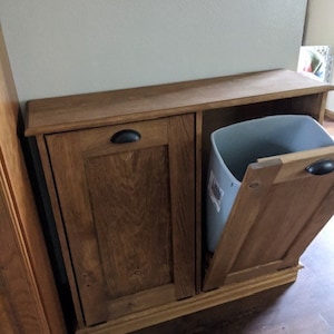 K&ouml;nnte beinhalten: Ein Holzschrank mit zwei T&uuml;ren und einem herausziehbaren M&uuml;lleimer. Der Schrank ist in einem dunklen Braunton gebeizt und hat ein schlichtes Design. Der M&uuml;lleimer ist aus grauem Kunststoff und ist teilweise sichtbar.