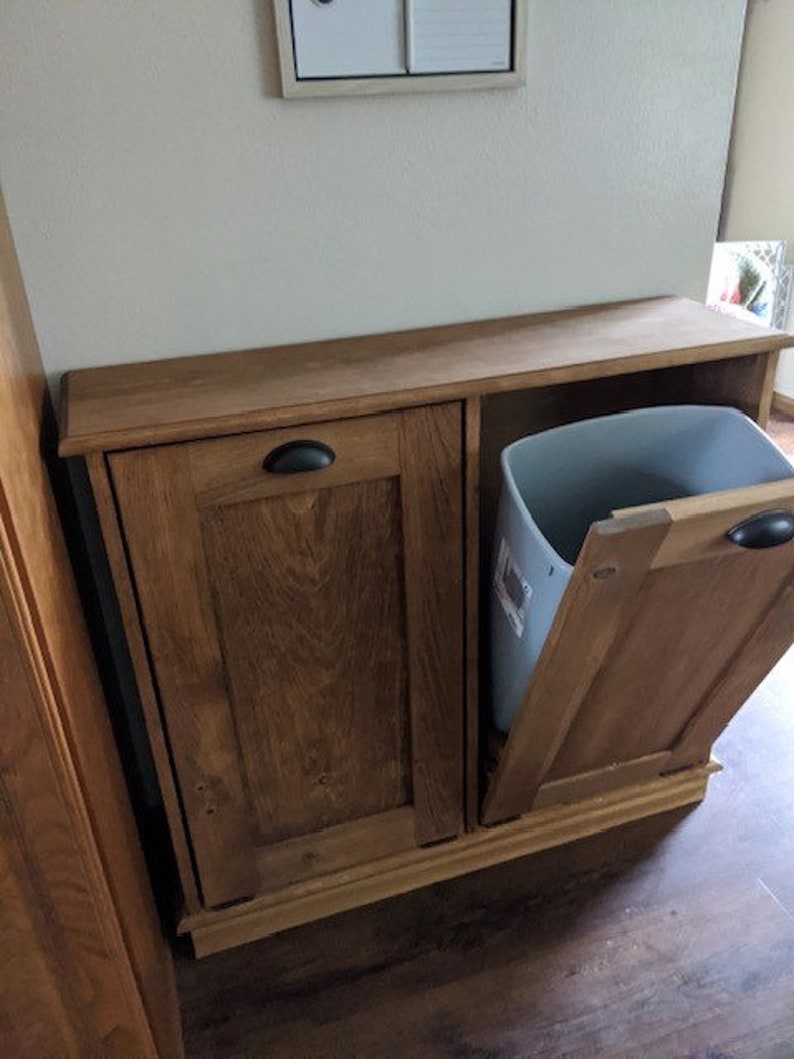 K&ouml;nnte beinhalten: Ein Holzschrank mit zwei T&uuml;ren und einem herausziehbaren M&uuml;lleimer. Der Schrank ist in einem dunklen Braunton gebeizt und hat ein schlichtes Design. Der M&uuml;lleimer ist aus grauem Kunststoff und ist teilweise sichtbar.