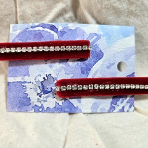 May include: Two red velvet hair clips with a row of clear rhinestones. The clips are displayed on a white card with a blue watercolor floral design.