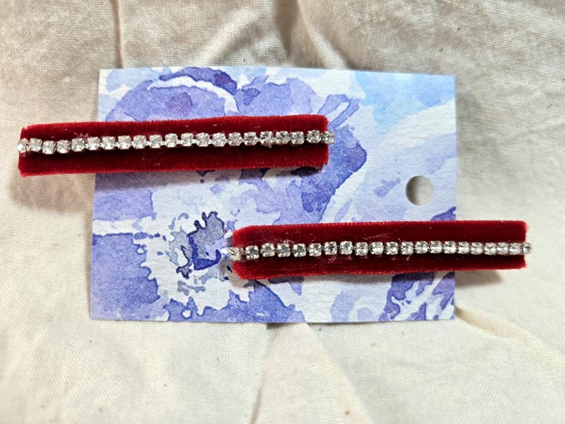 May include: Two red velvet hair clips with a row of clear rhinestones. The clips are displayed on a white card with a blue watercolor floral design.