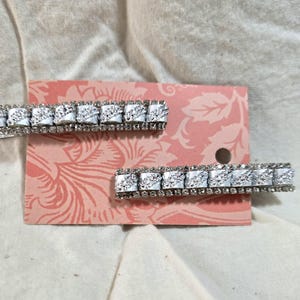 May include: Two silver hair clips with rows of clear rhinestones. The clips are on a pink and white floral patterned background.