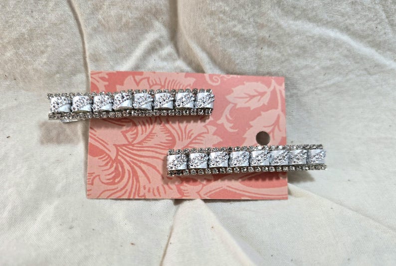 May include: Two silver hair clips with rows of clear rhinestones. The clips are on a pink and white floral patterned background.