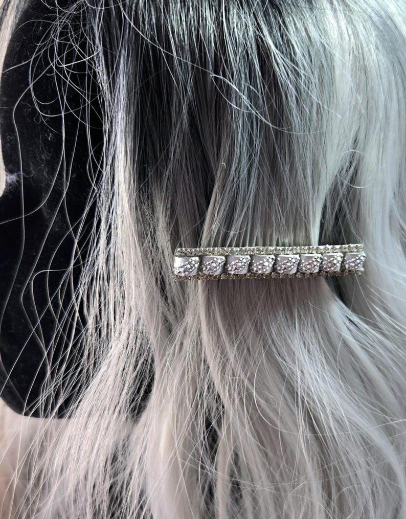 May include: A silver hair clip with a row of clear rhinestones. The clip is in the shape of a rectangle and has a textured surface.