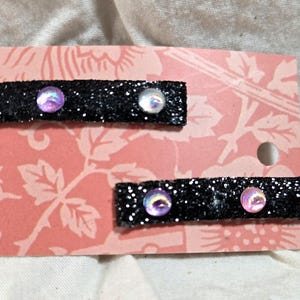May include: Two black glitter hair clips with iridescent gems. The clips are on a pink and white patterned background.