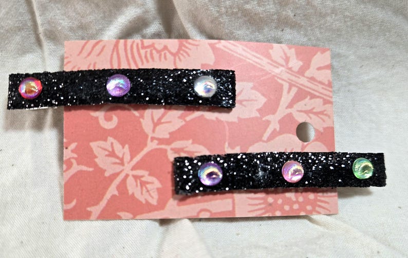 May include: Two black glitter hair clips with iridescent gems. The clips are on a pink and white patterned background.