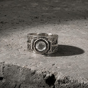 Peut inclure: Une large bague en argent avec une surface texturée et patinée, dotée d'un sertissage circulaire central. Le centre contient une pierre ronde, lisse, translucide et incolore. Le métal présente une finition oxydée sombre avec des détails.