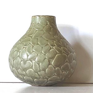 May include: A sage green ceramic vase with a rounded, bulbous body and a narrow neck. The exterior surface features a raised, textured pattern of irregular, pebble-like shapes that cover the entire body of the vessel in a repeating design.