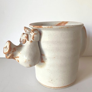May include: A handmade ceramic mug with a light off-white glaze and rustic orange-brown accents along the rim and base. A sculpted three-dimensional pig head protrudes from the side of the mug, featuring a snout, ears, and eyes in the same finish.
