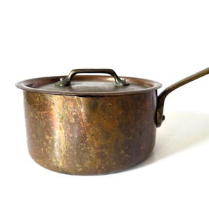 May include: A round copper saucepan with a weathered, mottled patina finish. The pot features a matching copper lid with a brass handle and a long, angled brass handle attached to the side. The surface shows visible signs of oxidation and age.