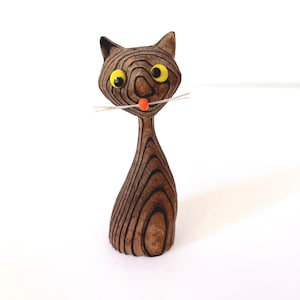 May include: Wooden cat figurine with a unique layered design. The cat has large yellow eyes, white whiskers, and a small orange nose. The figurine is brown with dark brown lines, and stands upright on a white surface.