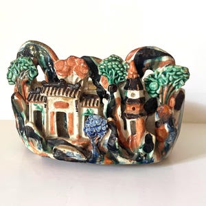 May include: A ceramic decorative piece featuring a sculpted landscape with a central building and a pagoda. The scene is painted in shades of green, brown, blue, and cream, depicting stylized trees, rock formations, and architectural structures.