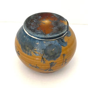 May include: A round ceramic jar with a lid, featuring a warm ochre-yellow base glaze. The surface is decorated with crystalline blue and dark grey patterns that resemble starbursts or floral shapes, distributed across the body and the lid.