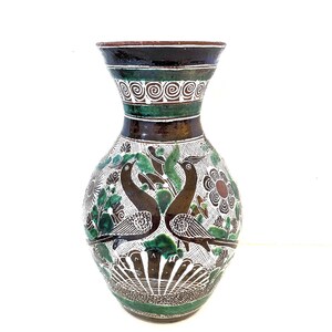 May include: A handcrafted ceramic vase with a wide base and a flared top. The vase features a dark brown rim and a green and white decorative band. The body is adorned with a detailed design of two birds, flowers, and foliage in shades of green, brown, and white.