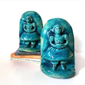 May include: A pair of matching teal ceramic bookends, each featuring a relief sculpture of a figure wearing a dress and a cap. The figures are centered within an arched frame. The bookends are positioned on top of a stack of three books.