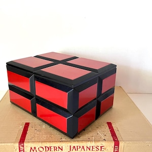 May include: A red and black geometric box with a lid. The box is made of wood and has a glossy finish. The box is sitting on a cardboard box with the words "Modern Japanese" printed on it.