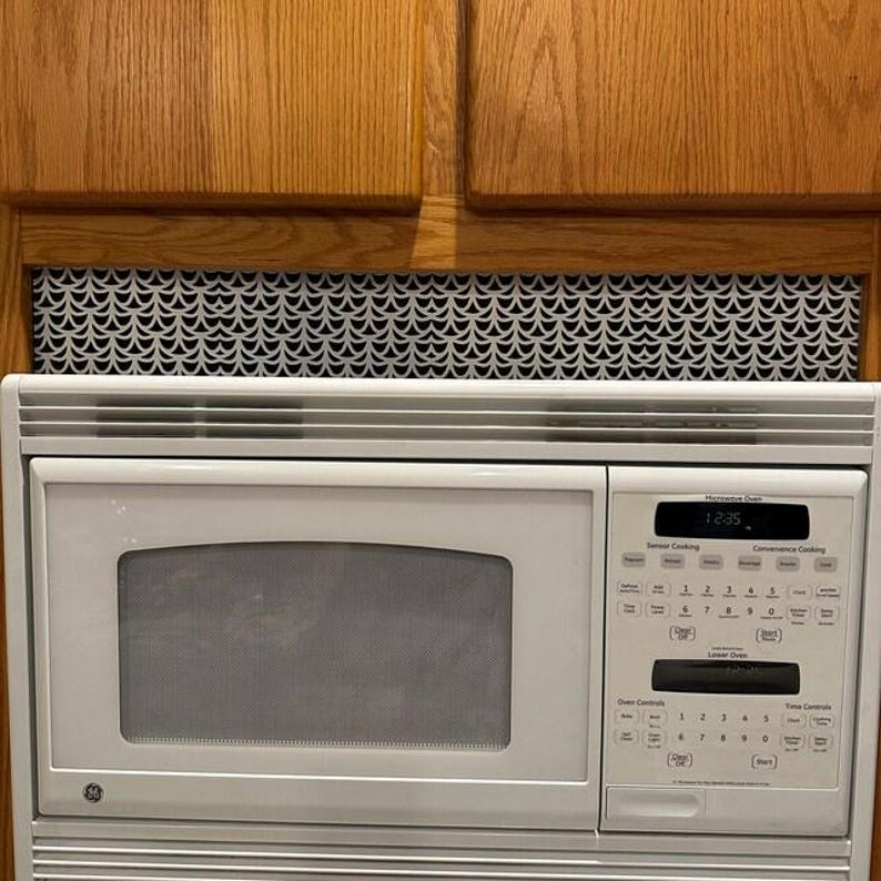 Puede incluir: Un horno microondas GE blanco con una pantalla de reloj digital que muestra las 12:35. El microondas est&aacute; integrado en un armario de madera con una rejilla de ventilaci&oacute;n decorativa con estampado en blanco y negro encima.