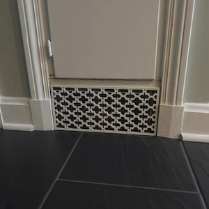 Puede incluir: Una rejilla de ventilaci&oacute;n de metal blanco con un patr&oacute;n geom&eacute;trico decorativo est&aacute; instalada cerca del suelo. La rejilla es rectangular y tiene un ojo de cerradura. La pared y los adornos circundantes est&aacute;n pintados de blanco.