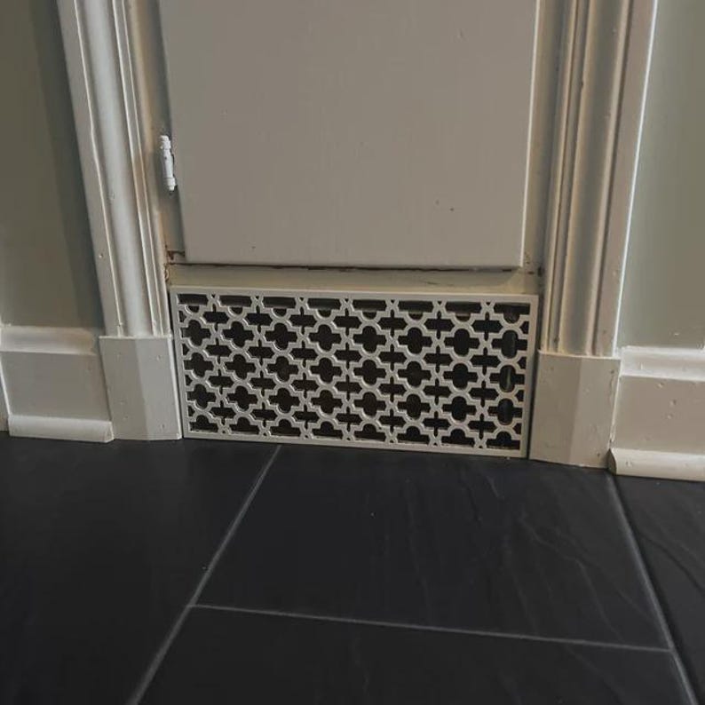 Puede incluir: Una rejilla de ventilaci&oacute;n de metal blanco con un patr&oacute;n geom&eacute;trico decorativo est&aacute; instalada cerca del suelo. La rejilla es rectangular y tiene un ojo de cerradura. La pared y los adornos circundantes est&aacute;n pintados de blanco.
