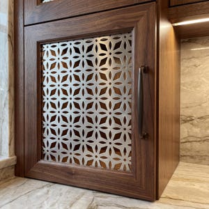 May include: A wooden cabinet door with a decorative metal screen featuring a repeating floral pattern. The cabinet has a dark brown finish and a long, bronze-colored handle. The screen is a light silver color.