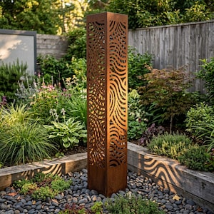 May include: A tall, square, rust-colored metal garden pillar featuring intricate, laser-cut wavy patterns on all four sides. The structure has a weathered, oxidized finish and stands upright, casting distinct wave-shaped shadows on the ground.