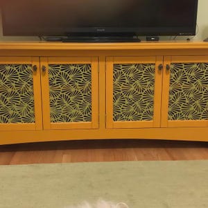 Puede incluir: Un mueble amarillo mostaza con cuatro puertas. Cada puerta presenta un patr&oacute;n decorativo en forma de abanico en un color verde m&aacute;s claro. El mueble tiene herrajes oscuros y est&aacute; debajo de un televisor negro.