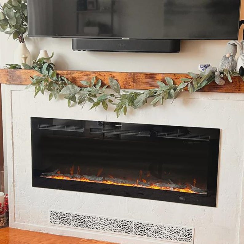 Puede incluir: Una chimenea el&eacute;ctrica moderna con una parte frontal de vidrio negro, que muestra un efecto de llama simulada. La chimenea est&aacute; empotrada en una pared blanca texturizada, con una repisa de madera decorativa encima. Una guirnalda verde y un televisor est&aacute;n montados encima.