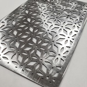 Puede incluir: Una l&aacute;mina met&aacute;lica rectangular con un patr&oacute;n floral recortado. La l&aacute;mina tiene un acabado plateado cepillado y una serie de formas de flores y diamantes. Los bordes son ligeramente redondeados.