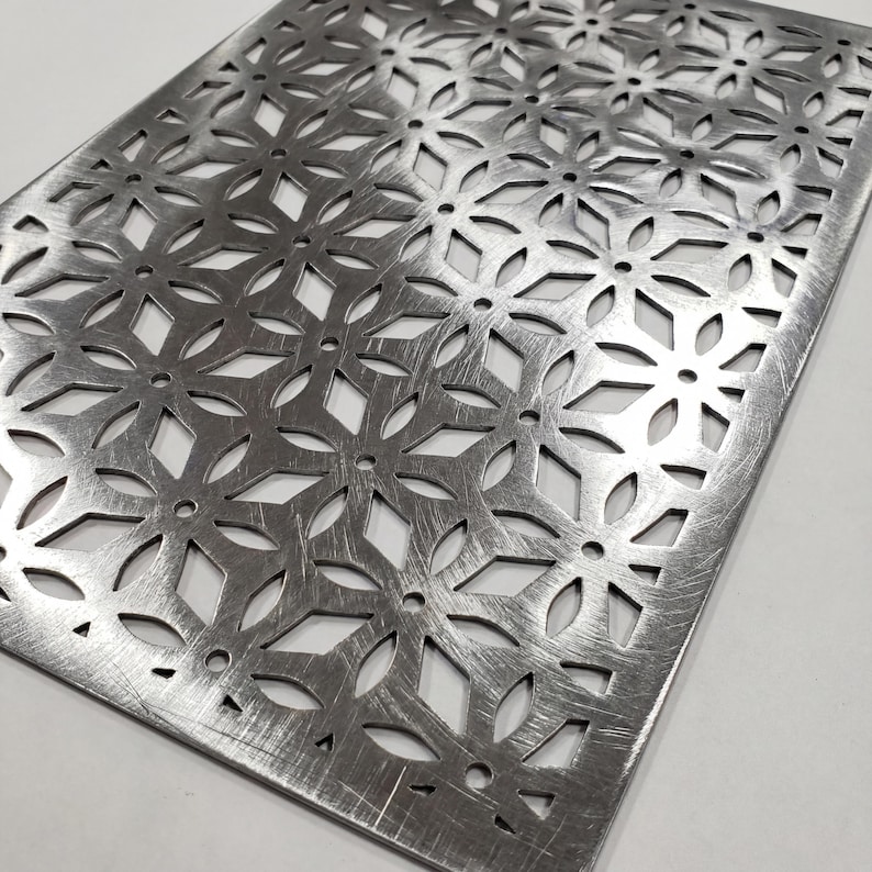 Puede incluir: Una l&aacute;mina met&aacute;lica rectangular con un patr&oacute;n floral recortado. La l&aacute;mina tiene un acabado plateado cepillado y una serie de formas de flores y diamantes. Los bordes son ligeramente redondeados.