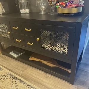 Puede incluir: Un aparador negro con herrajes dorados. El mueble tiene cajones y puertas con inserciones decorativas de malla met&aacute;lica. Una balda inferior contiene tablas de cortar de madera. El mueble est&aacute; sobre un suelo de madera.