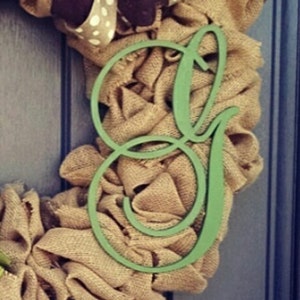 Può includere: Una ghirlanda di juta con la lettera "G" in corsivo verde chiaro al centro. La ghirlanda ha un aspetto naturale e rustico, con una combinazione di colori marrone e beige. Un fiocco marrone e bianco a pois è visibile in alto.